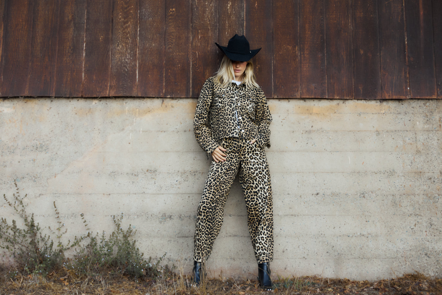 Person wearing a leopard print outfit and black hat against a rustic wall.