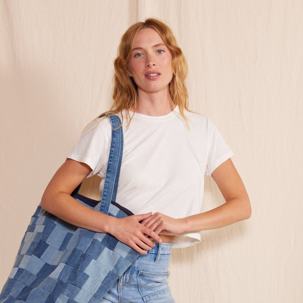 Woman wearing a white t-shirt and blue jeans holding a denim bag against a beige curtain background