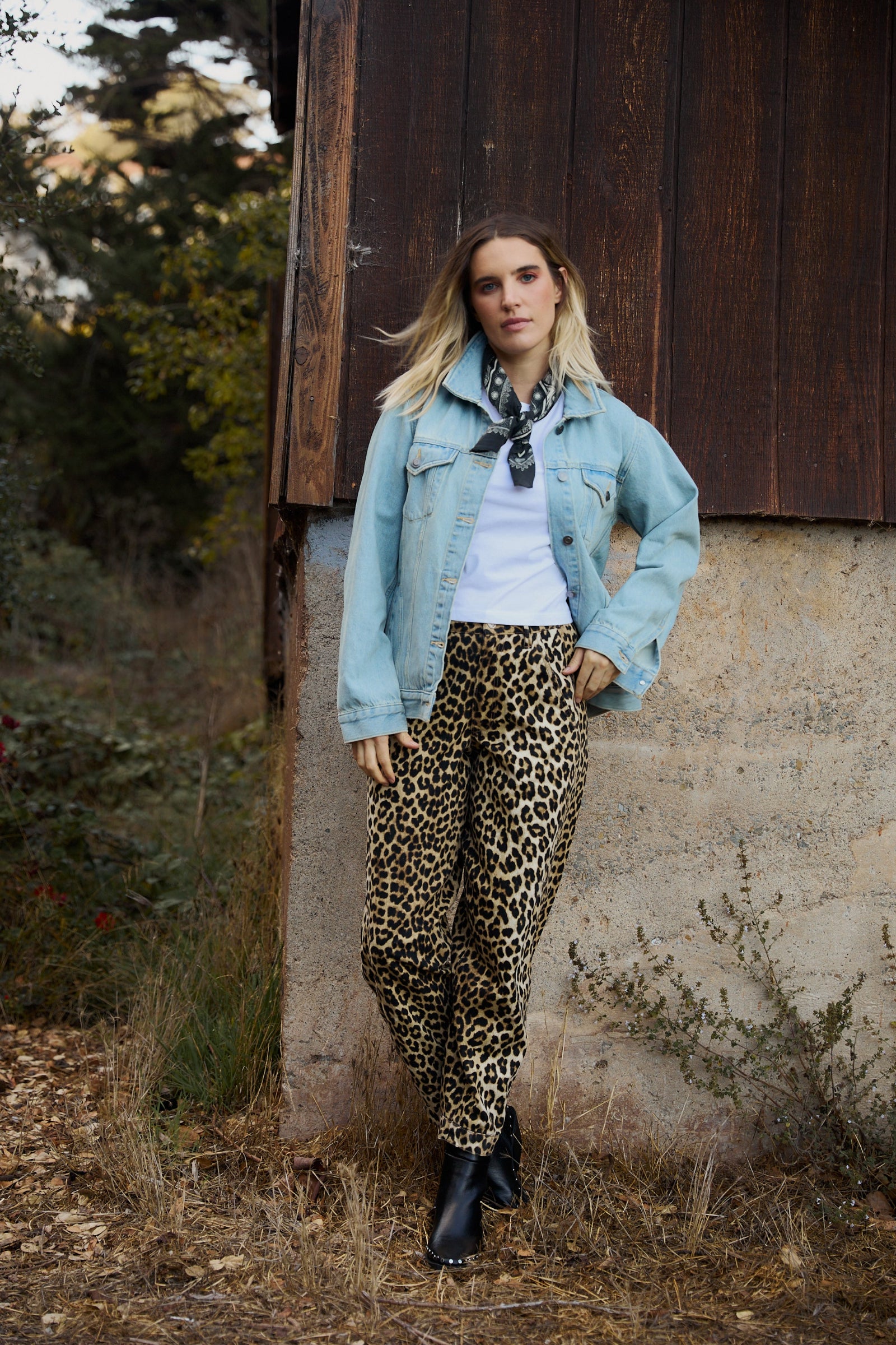 Woman wearing a light blue denim jacket, white shirt, and leopard print pants standing against a rustic wooden wall.